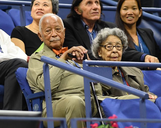 David Dinkins, wearing a khaki green outfit with orange bow tie and holding a cane sits in the stands with wife Joyce